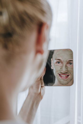Happy young make-up artist with facial mask looking at mirror