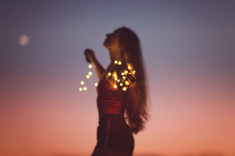 Side view of woman holding illuminated string lights against orange sky