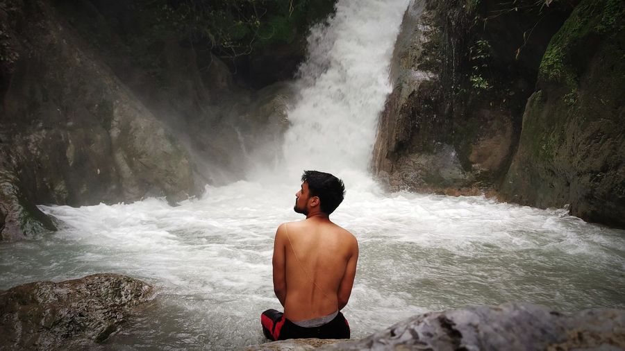 Rear view of shirtless man sitting against waterfall