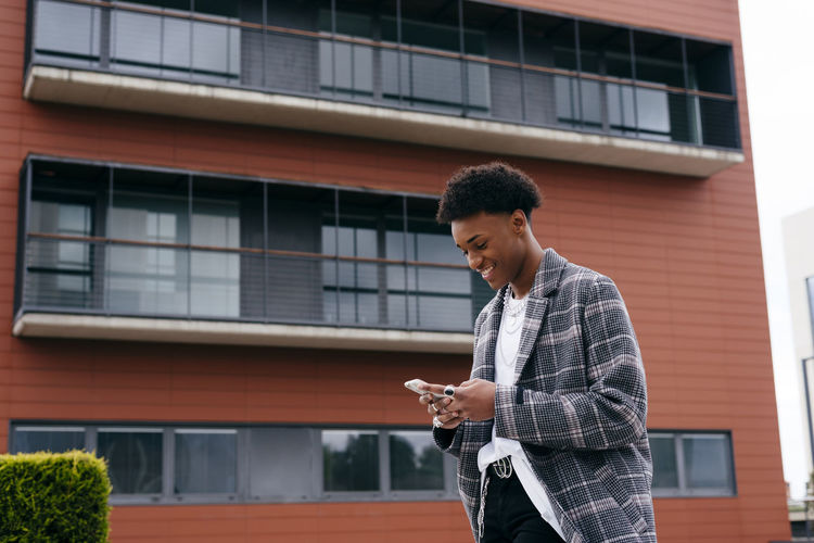 From below side view of happy young african american male student in stylish outfit browsing smartphone while standing on city street near modern building