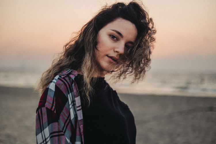 Portrait of beautiful young woman standing by sea against sky during sunset