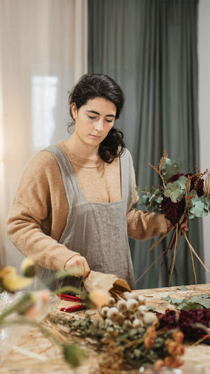 Diligent adult woman in casual clothes and gray apron making beautiful flower composition with dry green and burgundy plants in combination with brown stems while standing at table at home