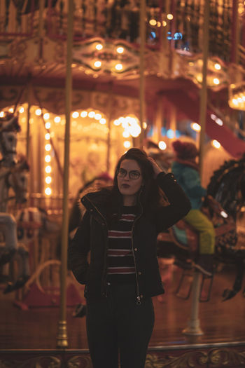 Portrait of young woman standing against illuminated carousel at night