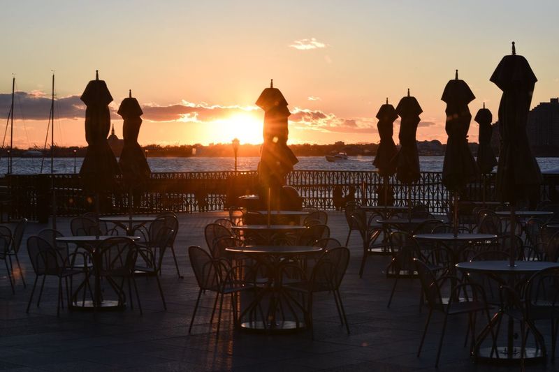 Empty chairs and tables by closed parasols at outdoor cafe during sunset