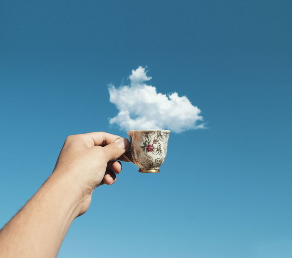 Optical illusion of cropped hand holding cup with cloud against blue sky