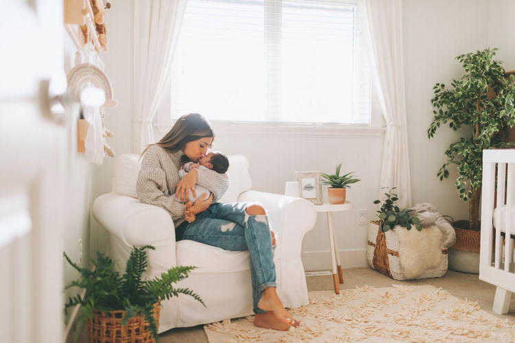 Full length of mother embracing baby while sitting on sofa at home