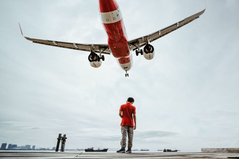 Rear view of boy standing on pier against airplane flying in sky