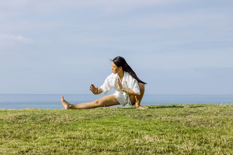 Young ethnic female squatting in skandasana pose while practicing yoga on grass coast against sea under cloudy sky