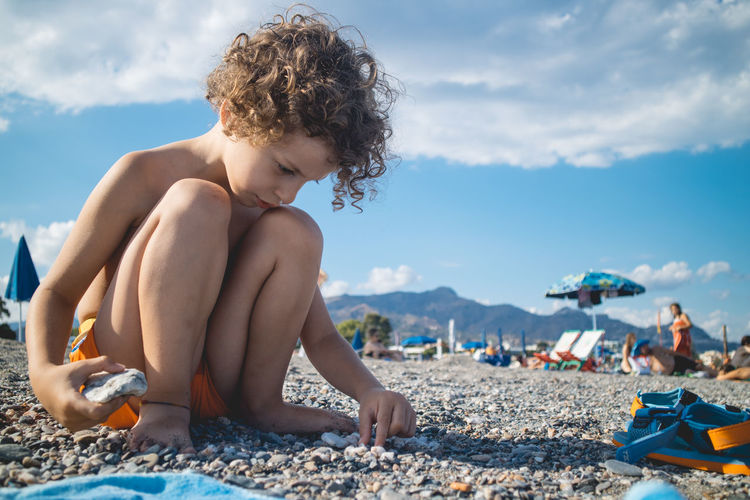 Shirtless boy playing with pebbles on beach against sky