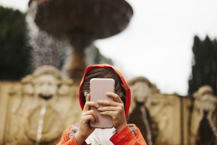 Boy wearing hooded shirt while using smart phone against fountain in city
