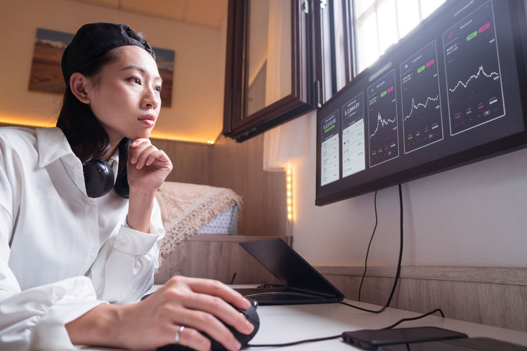 Side view of concentrated asian female working on computer with charts showing dynamic of changes in value of cryptocurrency at convenient workplace