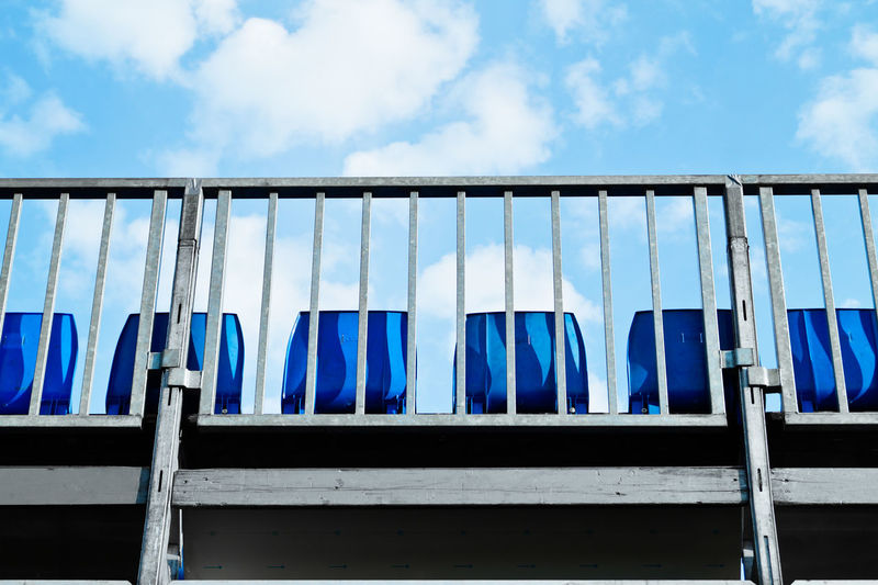 Low angle view of blue seats seen through fence against sky