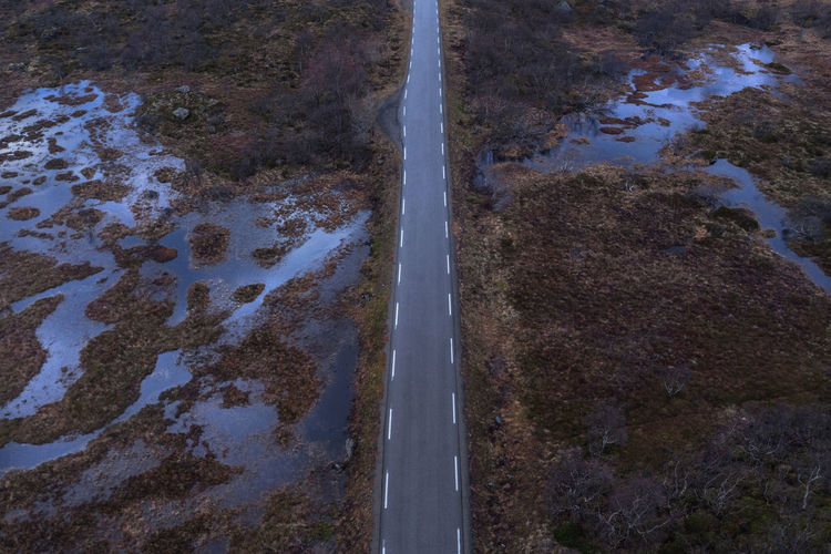 Drone view of long narrow asphalt roadway with making lines running between wetland with withered grass in nature of norway