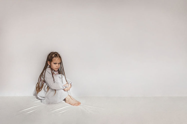Girl with braided hair sitting on bed against white wall