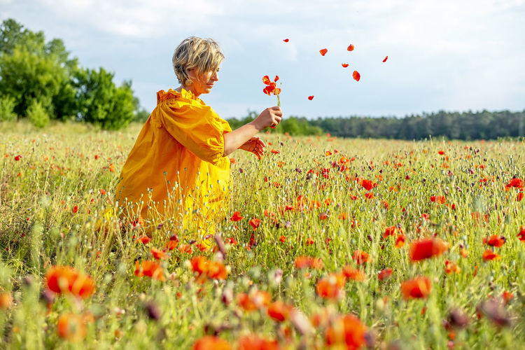 Side view of woman picking poppy flowers on field against sky