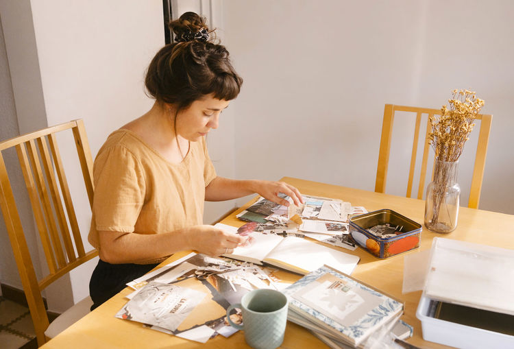 Freelancer arranging paper clippings in book on desk at home