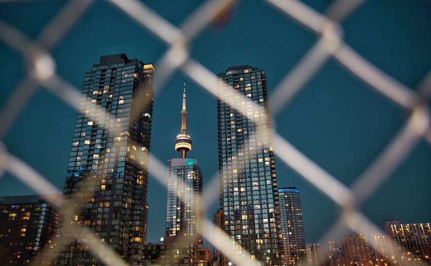 Low angle view of illuminated buildings seen through fence against sky