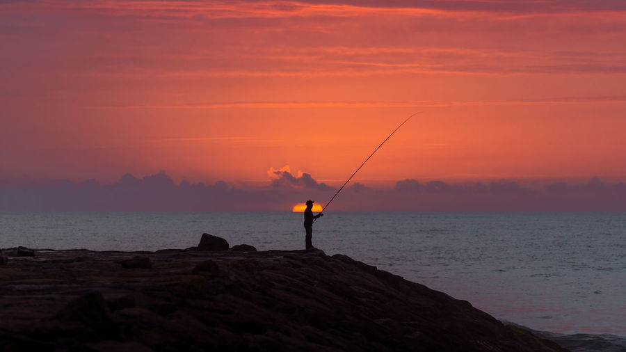 Silhouette man fishing by sea against orange sky during sunset