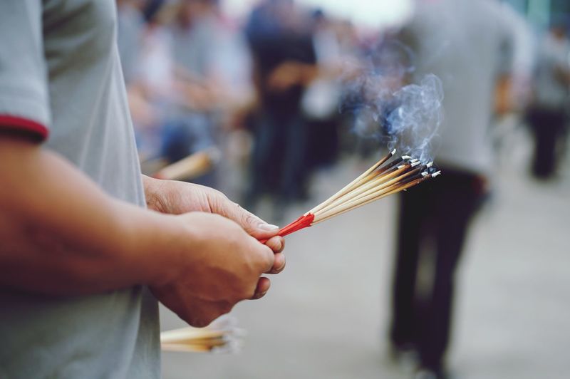 Close-up midsection of hands holding illuminated incense sticks in city