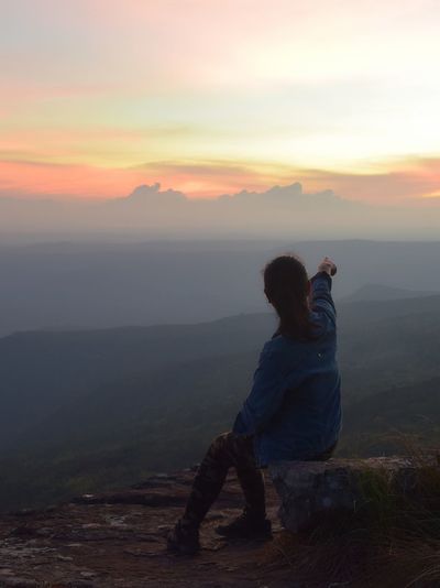 Rear view of woman sitting on mountain against sky at sunset
