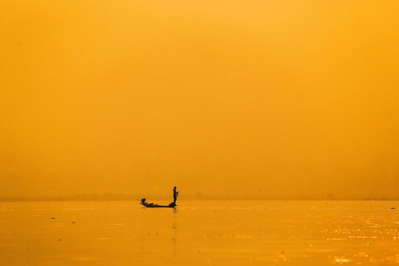 Silhouette fisherman on boat in sea against yellow sky during sunrise