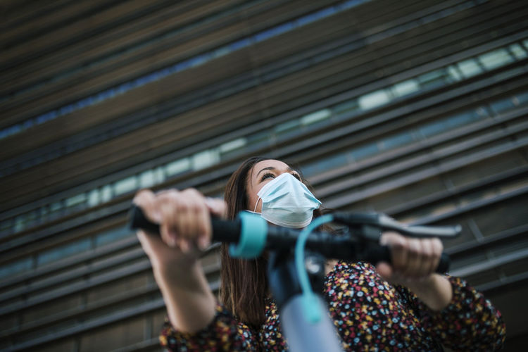 Mature woman in protective face mask with electric scooter against building during coronavirus crisis