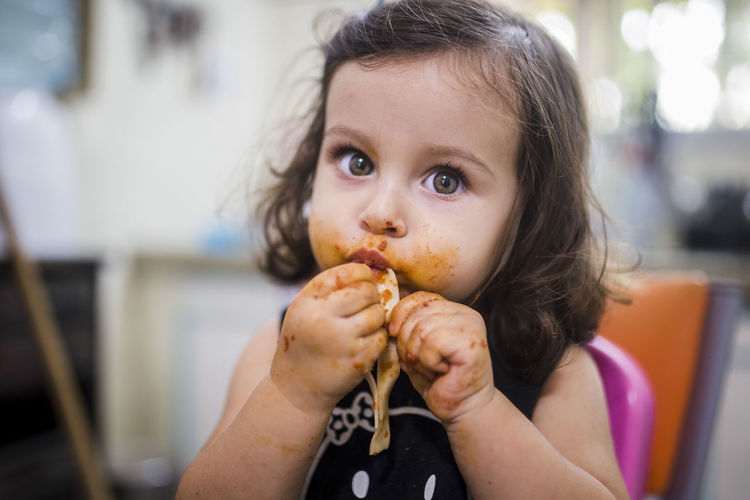 Close-up portrait of cute messy baby girl eating food while sitting at home