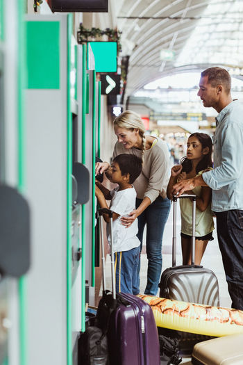 Mother assisting son in buying ticket while family standing with luggage on railroad station platform