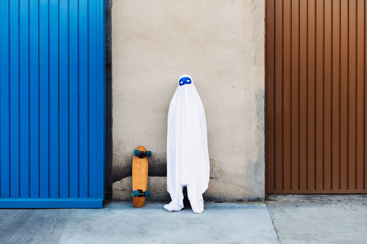 Unrecognizable child wearing mask and white ghost costume standing near skateboard in city during halloween and looking at camera