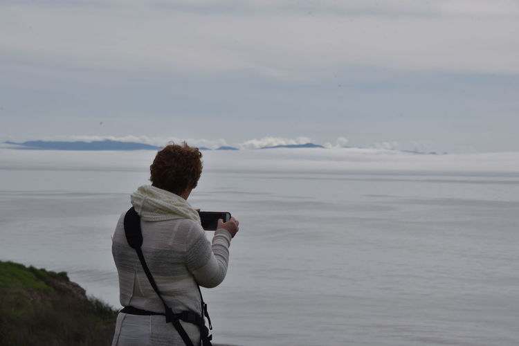 Rear view of woman photographing fog through smart phone against sky