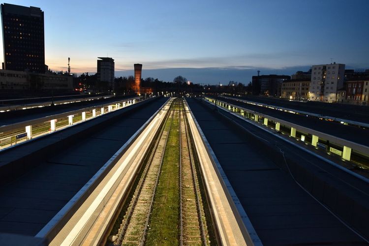 Railroad tracks by illuminated buildings against sky at dusk
