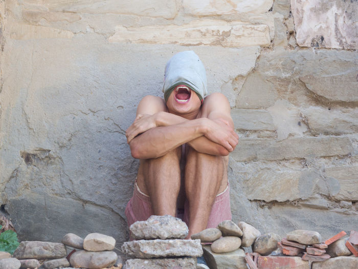 Shirtless young man screaming while sitting by wall