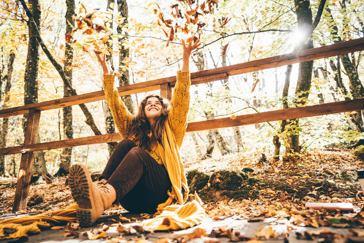 Positive curly haired woman in yellow sweater throwing dry leaves in picturesque autumn forest 