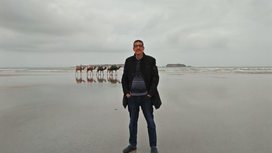 Portrait of man with hands in pockets standing at beach against cloudy sky