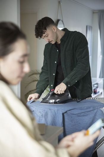 Young man ironing shirt while girlfriend using smart phone in living room
