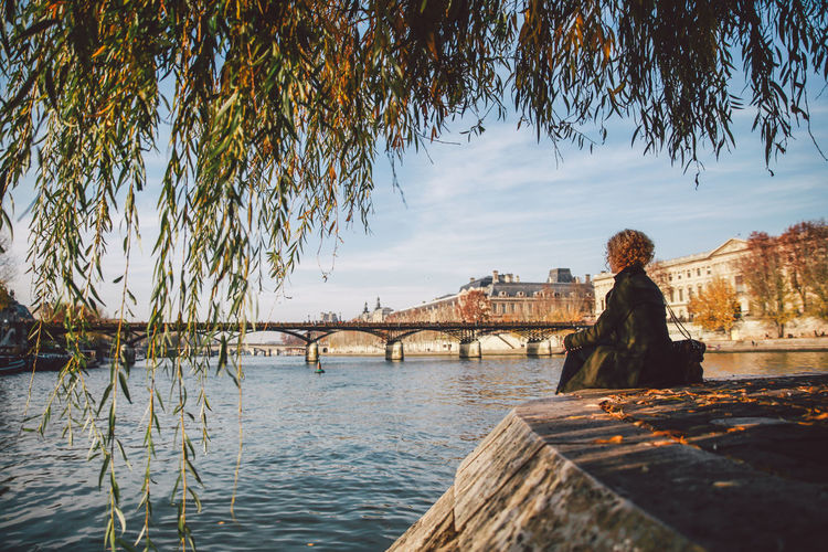 Young woman sitting at seine riverbank with tree leaves on foreground