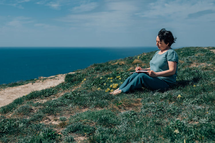Adult woman thoughtfully writes plans diary on a sunny day on a mountain against backdrop of the sea