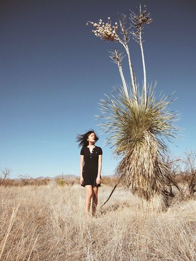 Woman jumping by plant on dry grassy field against clear blue sky