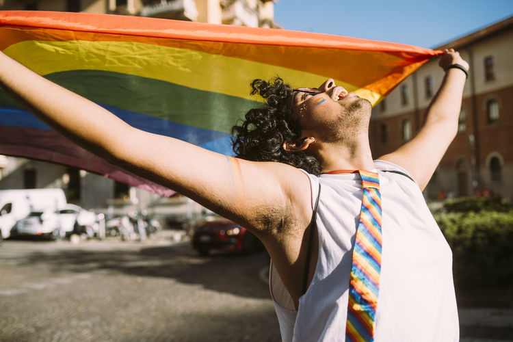 Young male activist looking up while holding rainbow flag on street