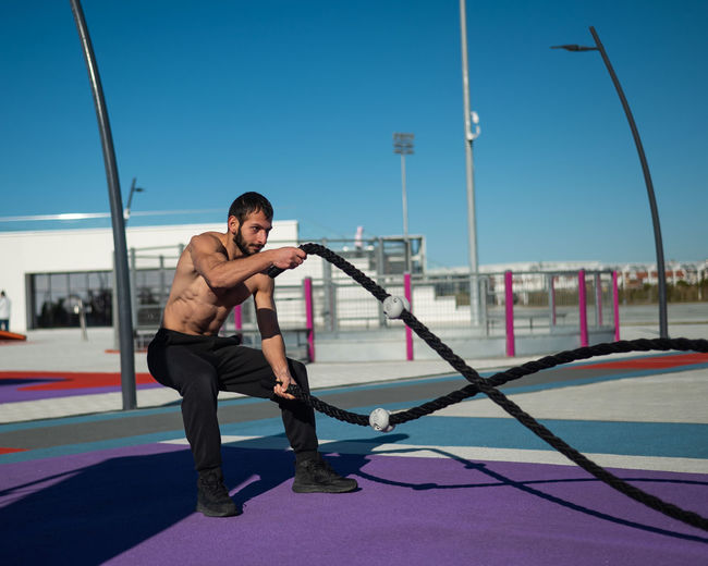 Shirtless man doing exercises with ropes outdoors. 