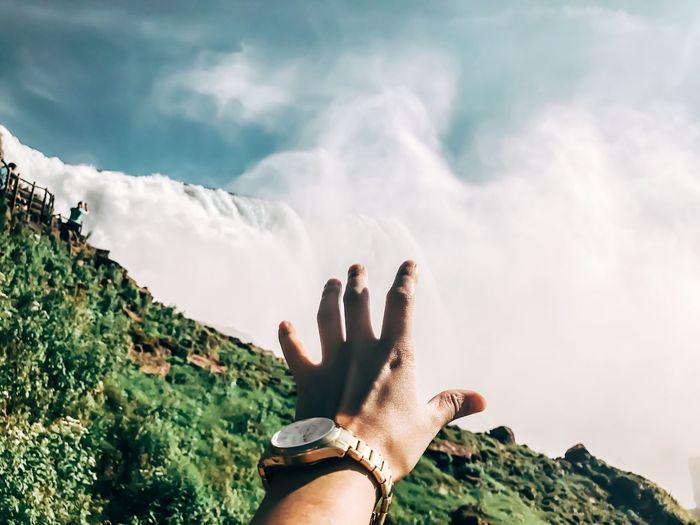 Cropped hand of man reaching waterfall against sky