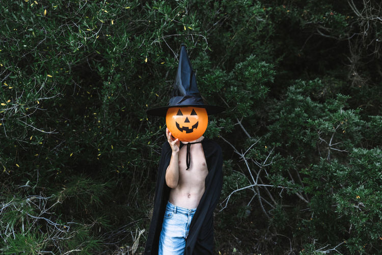Unrecognizable child in black halloween cloak covering face with air balloon in shape of pumpkin