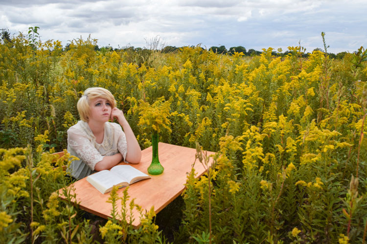 Thoughtful young woman sitting with book at table amidst plants on field