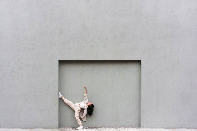 Creative cool female leaning on gray wall and dancing expressively in city street