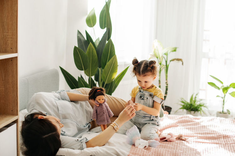A charming little girl plays with dolls with her mom on the bed during the day. motherhood, caring.