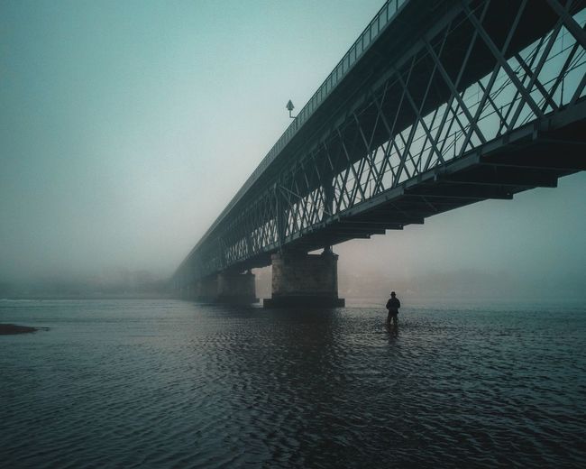 Silhouette person standing below bridge in lake against clear sky