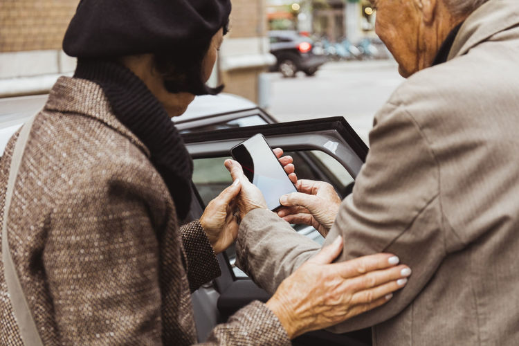 Senior couple using smart phone standing outside car in city during weekend trip