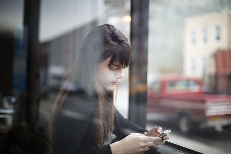 Close-up of young woman using smart phone seen through window