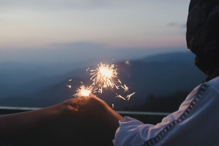 Woman with friend holding sparklers against sky during sunset