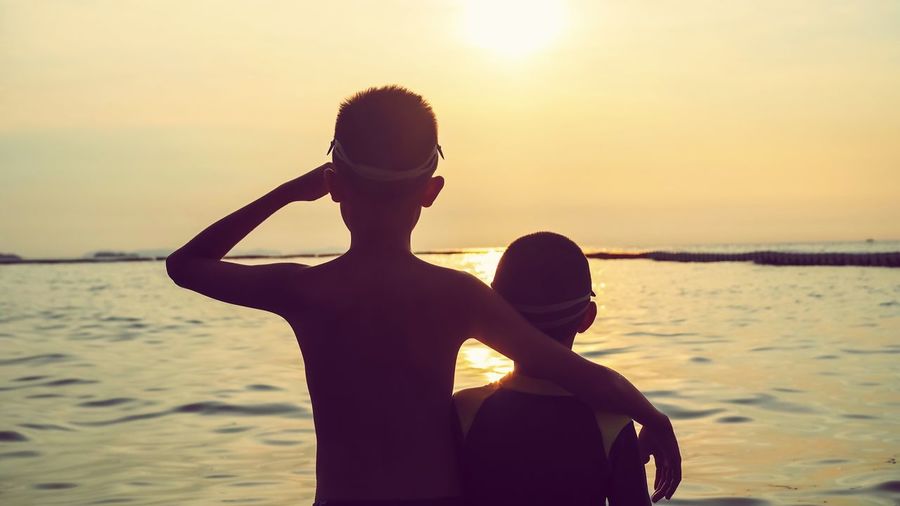 Rear view of boys looking at sea against sky during sunset